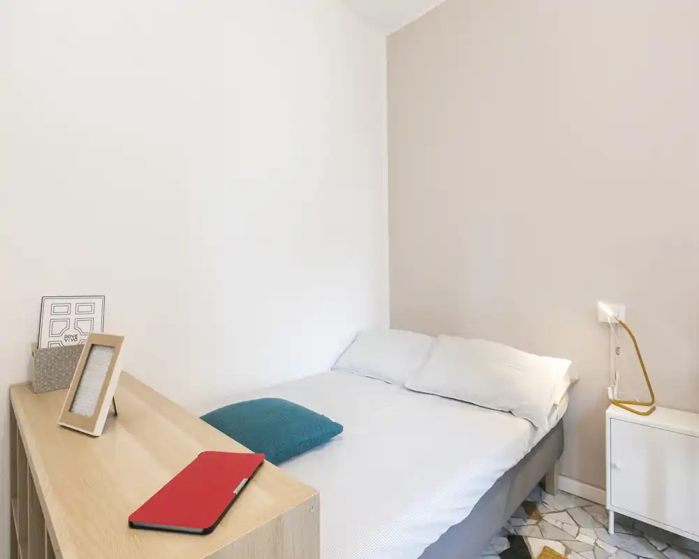 Dormitorio luminoso y minimalista con cama doble bien hecha, ropa de cama blanca, cojín turquesa y pequeña mesita; estilo sencillo y moderno, ideal para anuncios.