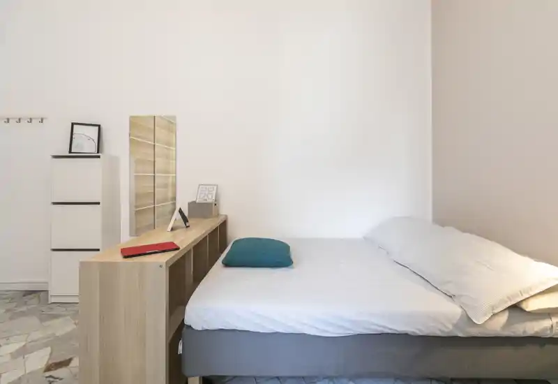 Bright, minimal bedroom with a neatly made double bed, a low shelving unit at the foot of the bed, and simple decorative accents for a calm, modern look.