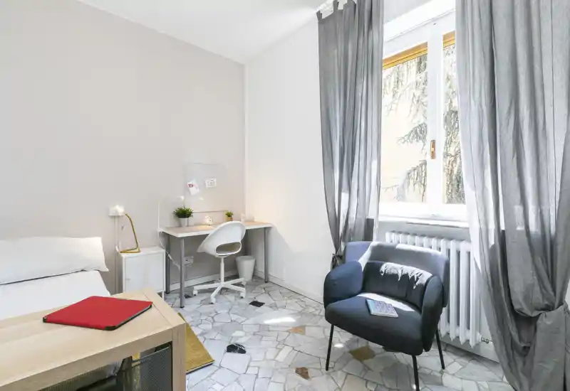 Dormitorio moderno y luminoso con cama cómoda, pequeño escritorio y un sillón acogedor junto a una gran ventana que deja entrar luz natural.