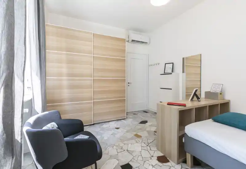 Bright modern bedroom with a single bed, a wooden storage unit at the foot, an armchair by the window and a large sliding-wardrobe. Clean white walls and natural light create a fresh, minimalist feel.
