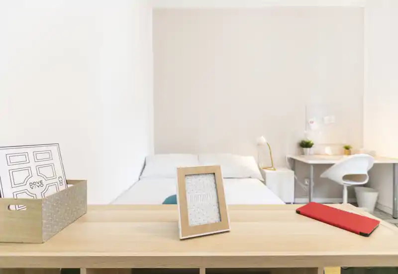 Dormitorio luminoso y minimalista con cama centrada frente a una pared clara, escritorio sencillo y detalles decorativos ordenados.
