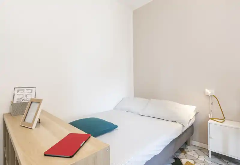 Dormitorio luminoso y minimalista con cama doble bien hecha, ropa de cama blanca, cojín turquesa y pequeña mesita; estilo sencillo y moderno, ideal para anuncios.