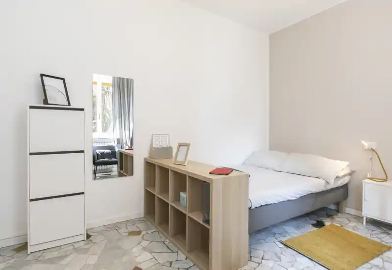 Dormitorio luminoso y minimalista con cama doble, mesita y estantería divisoria: tonos neutros y luz natural que hacen la estancia amplia y acogedora.