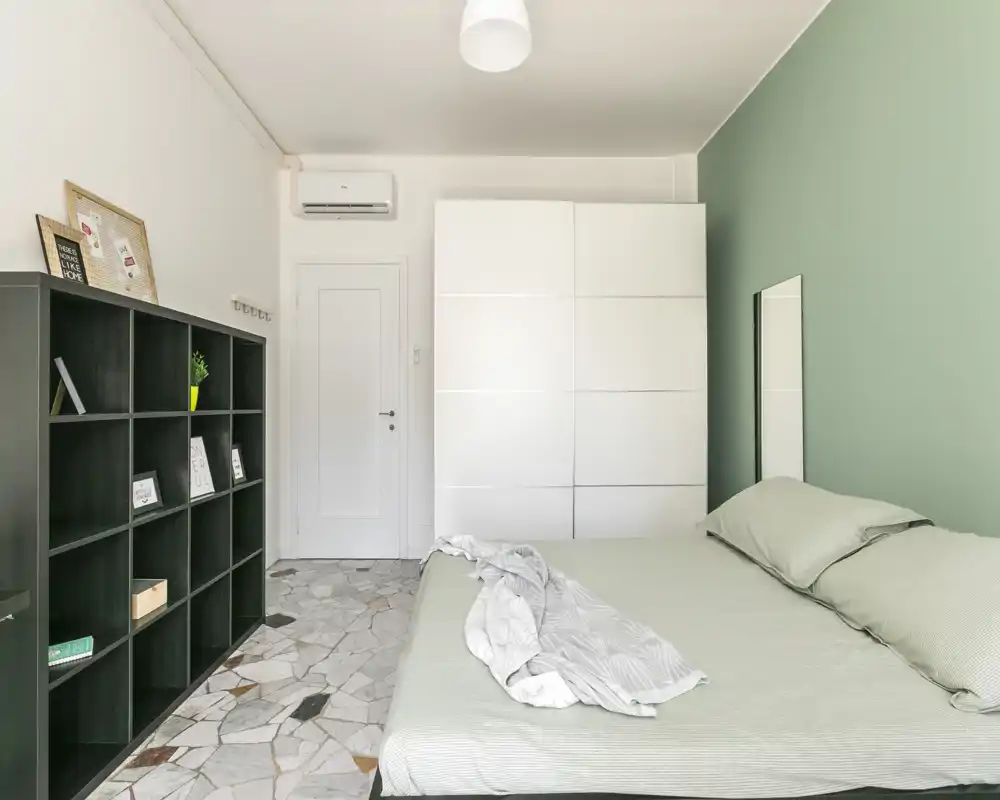 Bright, modern bedroom with a large double bed, soft green accent wall, white wardrobe and black shelving—minimalist and airy.