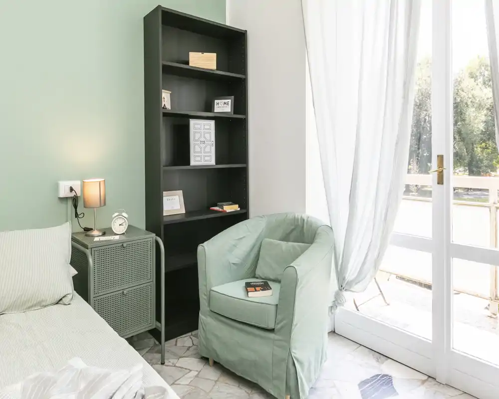 Bright, cozy bedroom corner with a comfortable single bed, soft armchair, bedside table and shelving; large glass door provides abundant natural light.