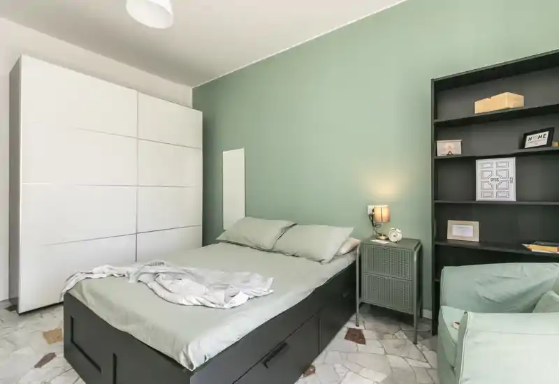 Bright, modern bedroom with a double bed, soft green accent wall, built-in storage and a cozy armchair — ideal for rental listings highlighting style and functionality.