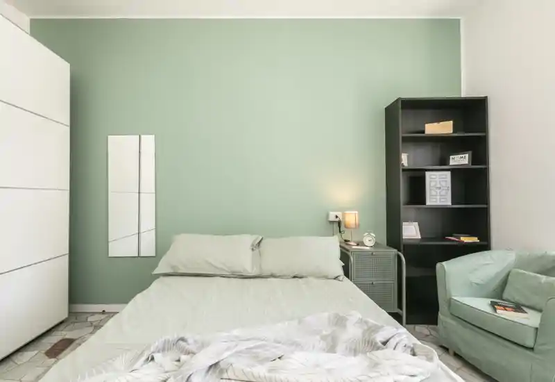 Bright, modern bedroom with a neatly made double bed against a soft green accent wall, a bedside lamp, shelving unit and a comfortable armchair — ideal for calm stays.