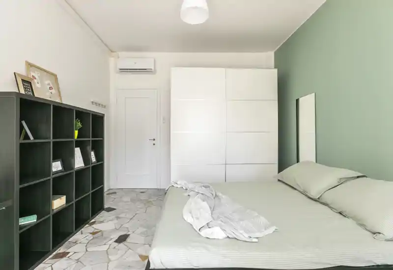 Bright, modern bedroom with a large double bed, soft green accent wall, white wardrobe and black shelving—minimalist and airy.