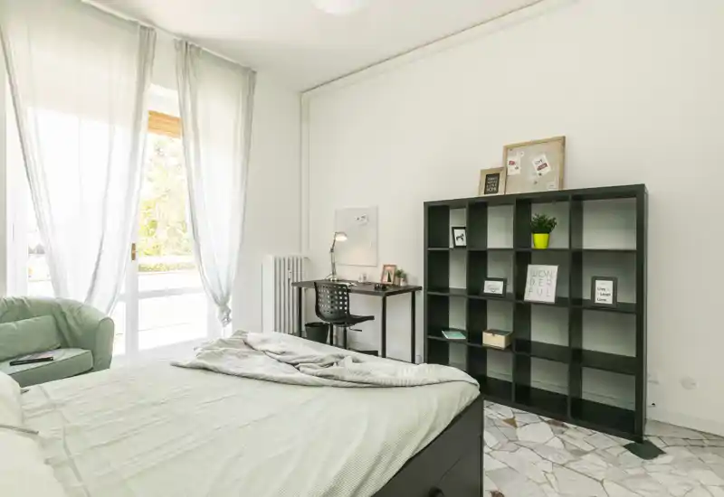Bright, modern bedroom with a neatly made double bed, a shelving unit and a small work desk by a large window letting in abundant natural light.