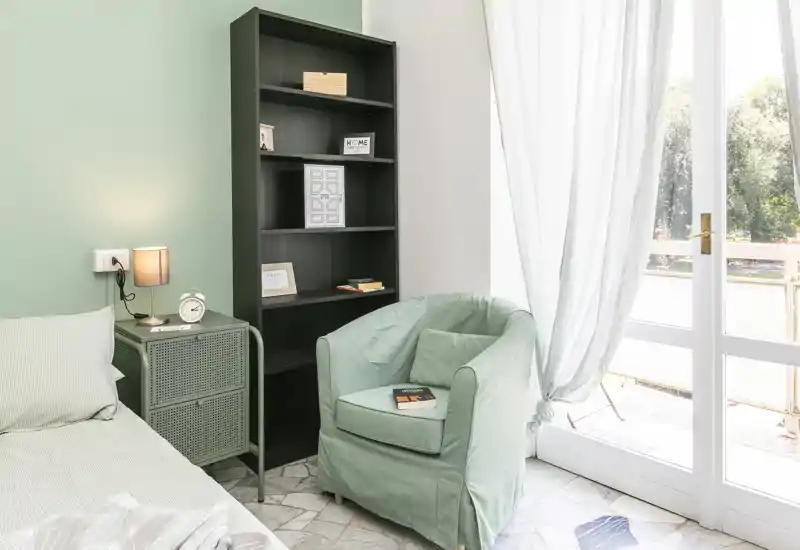 Bright, cozy bedroom corner with a comfortable single bed, soft armchair, bedside table and shelving; large glass door provides abundant natural light.