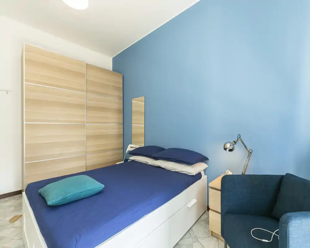 Dormitorio moderno y luminoso con cama doble vestida en tonos azul oscuro, sillón a juego y armario de madera clara. Pared azul de acento, mesita con lámpara y pequeño espejo: limpio, acogedor y bien iluminado.