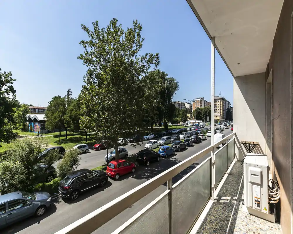 Balcón soleado con vistas a una calle arbolada y coches aparcados, suelo de baldosas y barandilla — ideal para mostrar el espacio exterior y la vista.