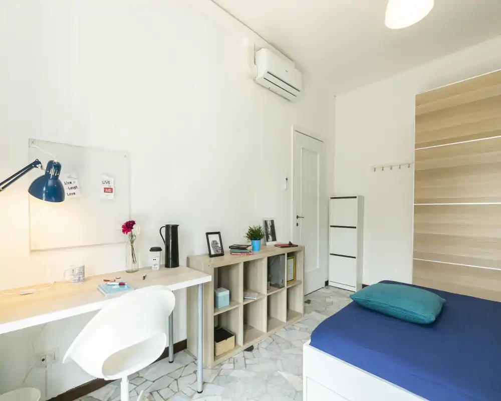 Dormitorio luminoso y minimalista con cama individual con ropa de cama azul, escritorio con lámpara y silla, y estantería para almacenamiento — ideal para estudiante o persona sola.
