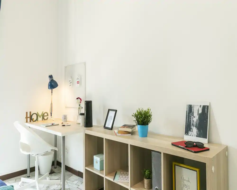 Espacio de trabajo luminoso y moderno con escritorio blanco, silla ergonómica y estantería abierta. La decoración con lámpara, planta y marcos crea un rincón de oficina en casa ordenado y acogedor.