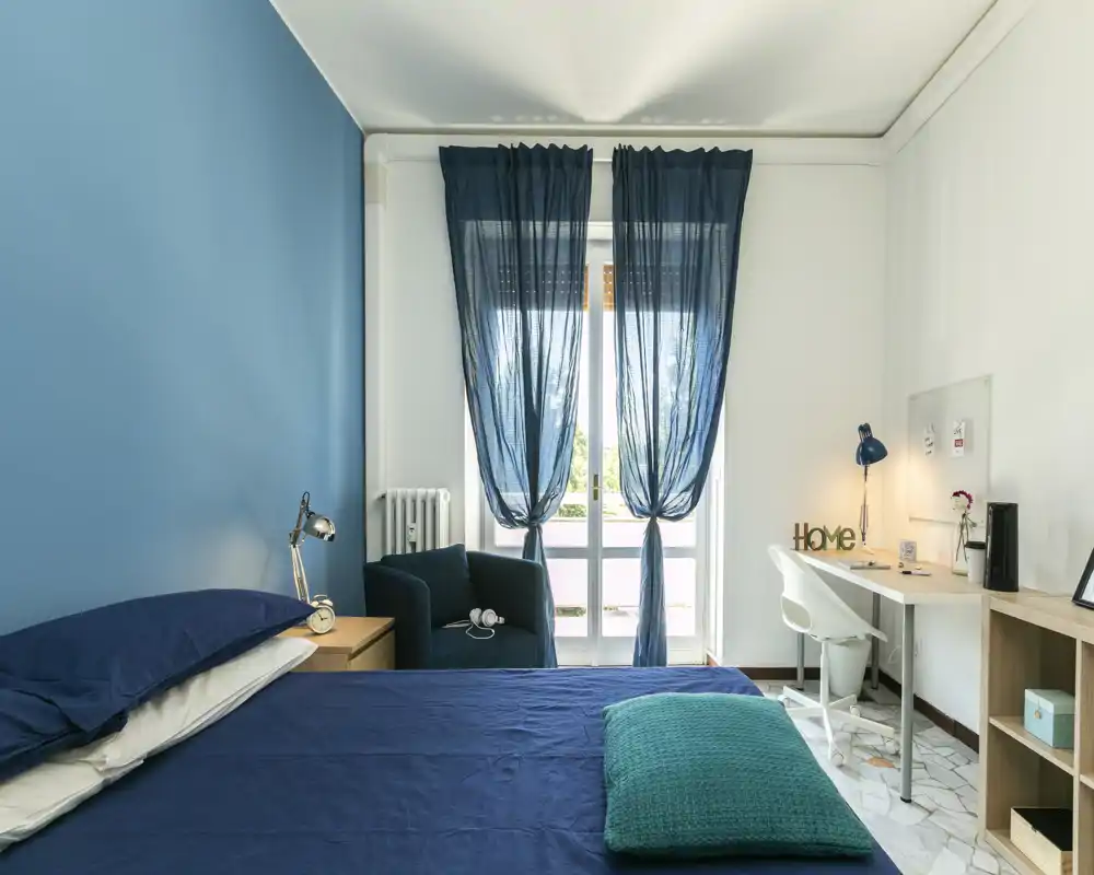 Dormitorio moderno y luminoso con pared azul, cama bien hecha y zona de estudio junto a la ventana. La luz natural llena la habitación; un sillón y una lámpara de noche crean un ambiente acogedor.