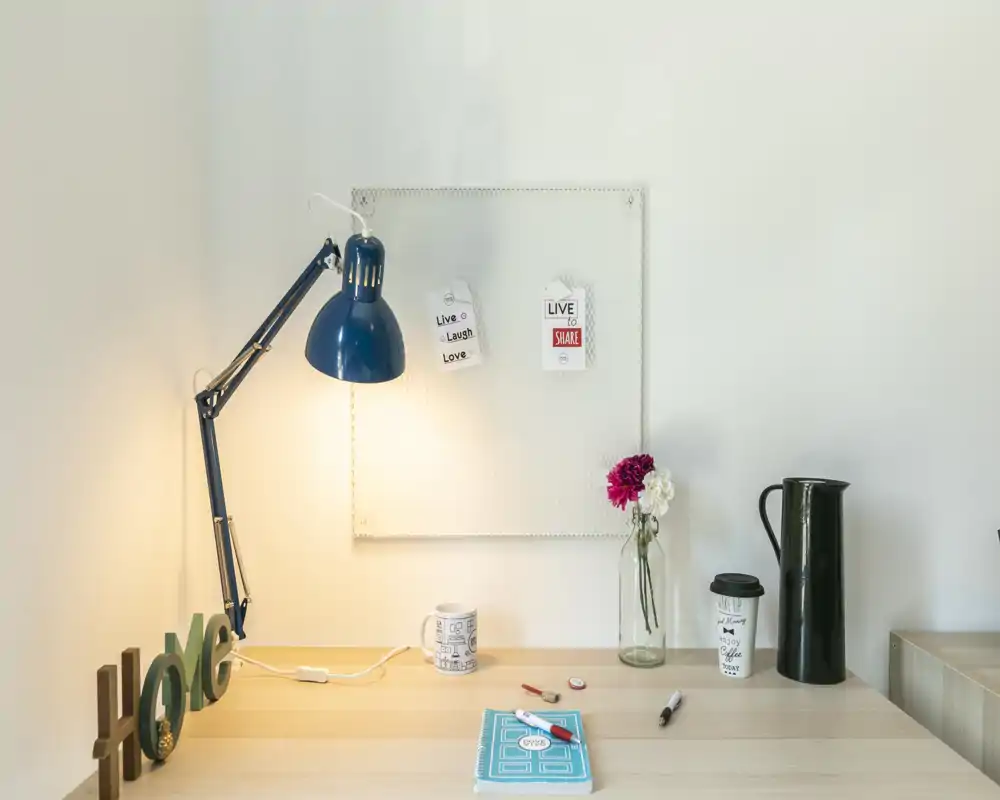 Escritorio ordenado con lámpara azul ajustable, cuaderno, tazas y un pequeño jarrón con flores: un espacio luminoso y minimalista ideal para trabajar o estudiar.