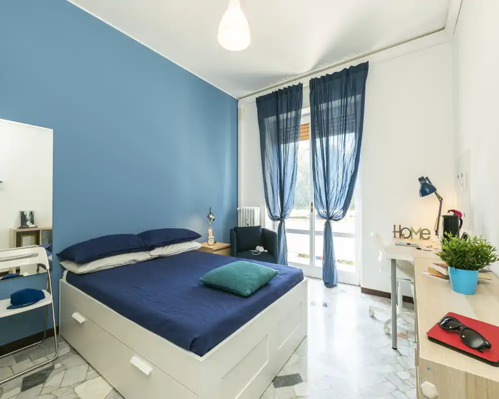 Dormitorio moderno y luminoso con cama de matrimonio, pared azul y cortinas transparentes que dan a un balcón; un escritorio y un sillón completan el ambiente acogedor.