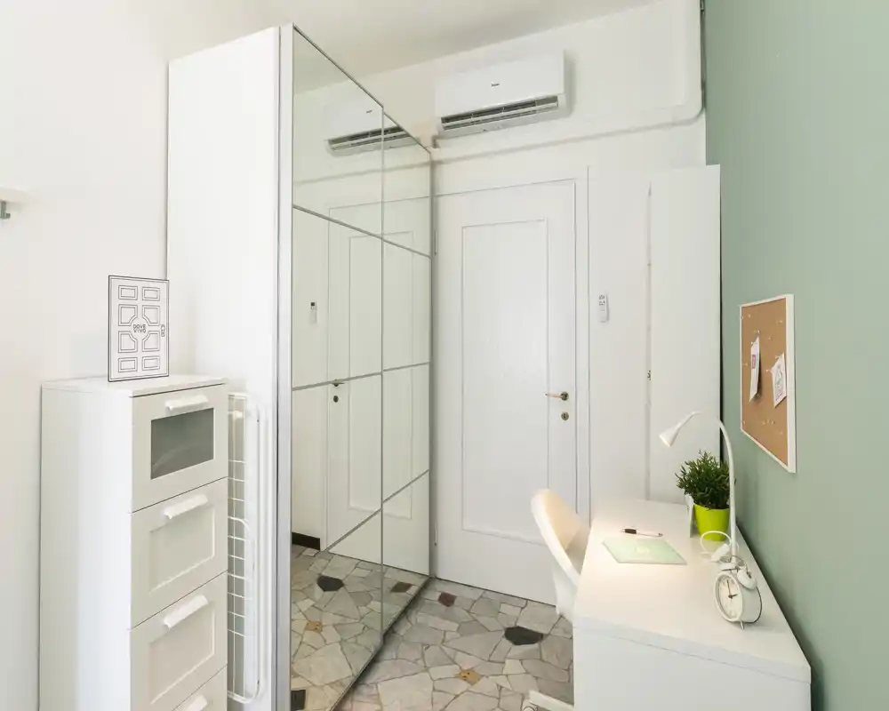 Rincón de dormitorio luminoso convertido en zona de trabajo con escritorio, silla, armario con espejo y cajonera; decoración minimalista con pared verde.