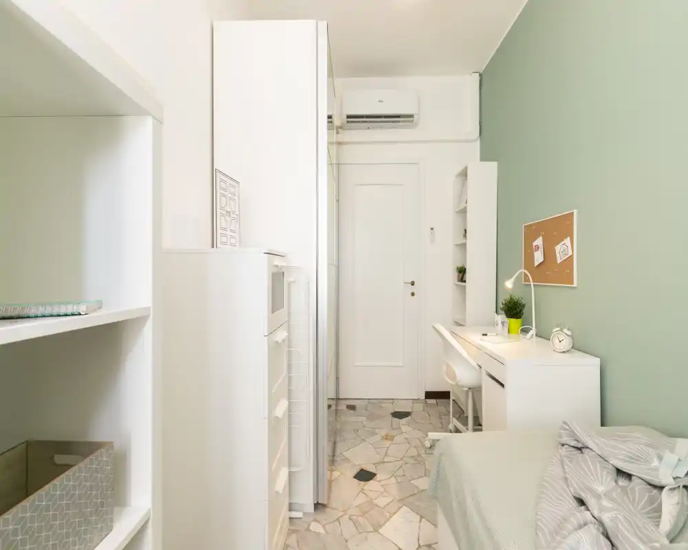 Dormitorio compacto y luminoso con cama individual, escritorio y armarios empotrados. Pared verde suave y muebles blancos que crean un ambiente moderno y tranquilo.