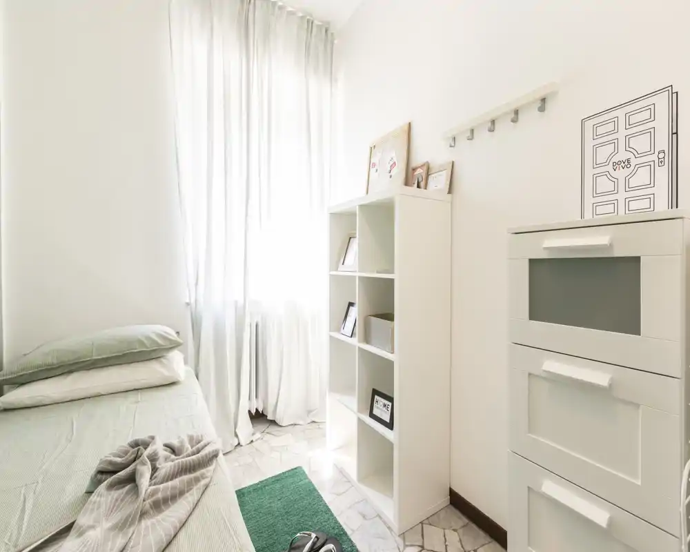 Habitación individual luminosa y minimalista con cama arreglada, estantería blanca y luz natural que entra por una ventana alta, creando un ambiente aireado.