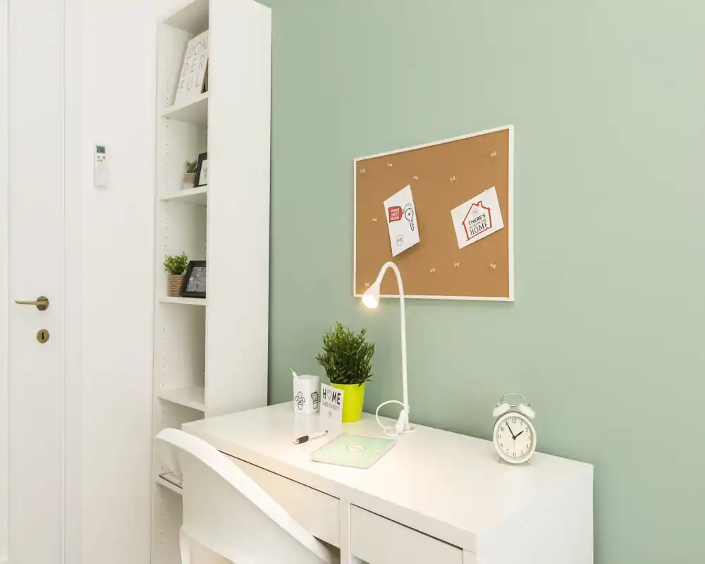 Área de trabajo luminosa y ordenada con escritorio blanco, silla, lámpara de escritorio, corcho para notas y planta decorativa — ideal para estudiar o teletrabajar.