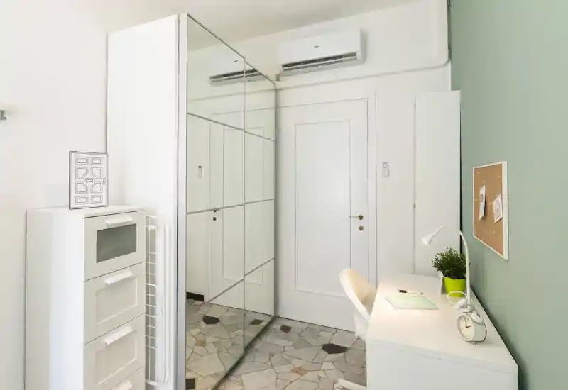 Small, bright bedroom corner styled as a workspace with a desk, chair, mirrored wardrobe and storage drawers; clean minimalist decor with a green accent wall and clock.