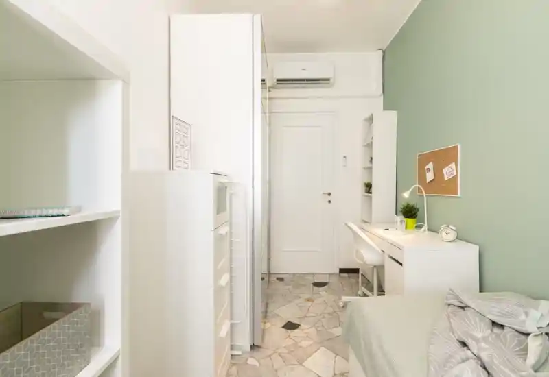 Dormitorio compacto y luminoso con cama individual, escritorio y armarios empotrados. Pared verde suave y muebles blancos que crean un ambiente moderno y tranquilo.