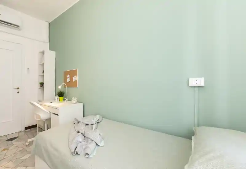 Dormitorio compacto y luminoso con cama individual frente a una pared verde menta, pequeño escritorio blanco con lámpara y silla, y decoración minimalista que crea un ambiente moderno y tranquilo.