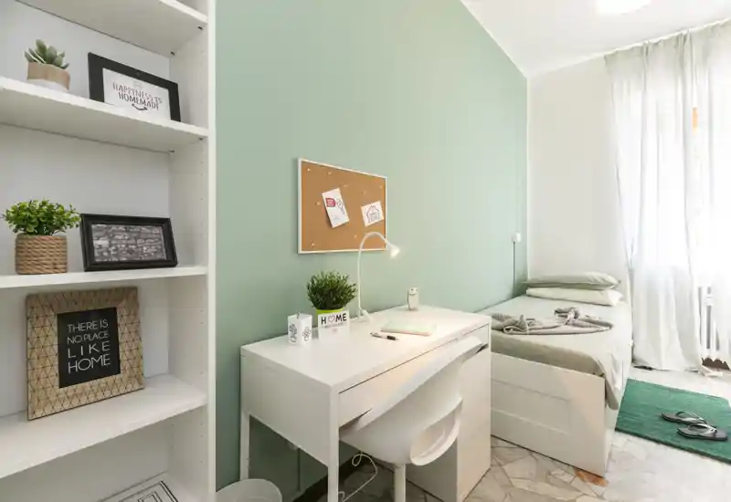 Dormitorio individual luminoso y cuidado con escritorio blanco, corcho en la pared y cama individual con ropa de cama suave. Pared verde claro, plantas y estantería crean un ambiente tranquilo y acogedor.