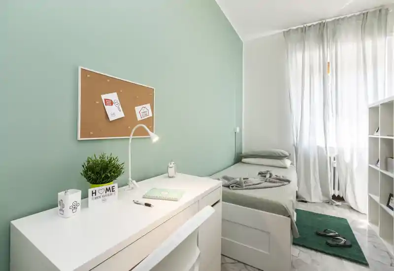 Habitación individual luminosa y ordenada con escritorio blanco, tablón de corcho y cama junto a una ventana con luz — ideal para estudiante o persona sola.