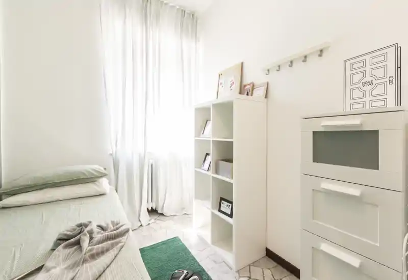 Habitación individual luminosa y minimalista con cama arreglada, estantería blanca y luz natural que entra por una ventana alta, creando un ambiente aireado.