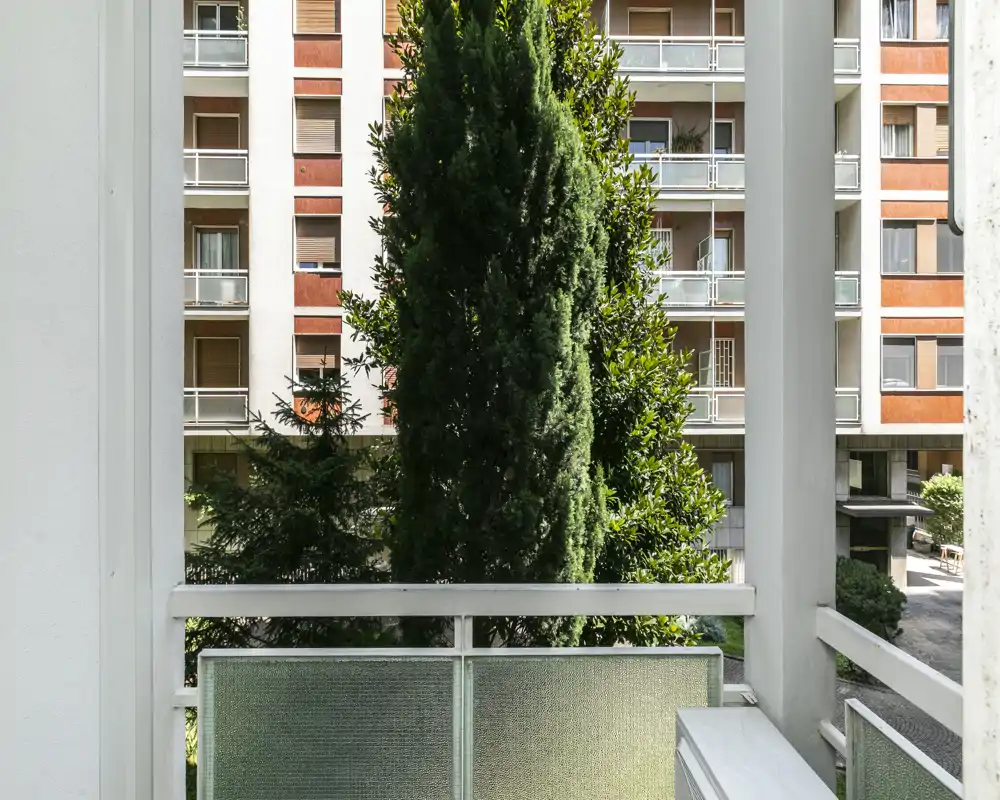 Pequeño balcón privado con vistas a un patio con árboles y fachadas de apartamentos vecinos, con barandilla de paneles de vidrio y buena luz natural.