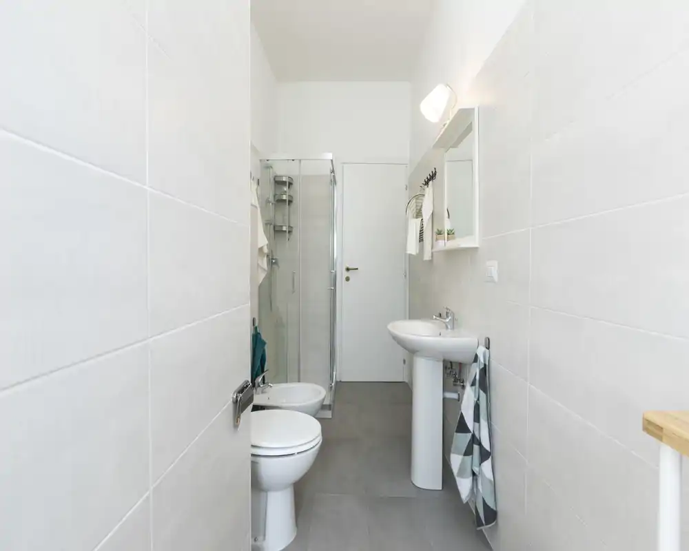 Baño moderno y luminoso con lavabo de pedestal, inodoro, bidé y ducha con mampara de cristal. Baldosas en tonos neutros y accesorios sencillos para un aspecto limpio y minimalista.