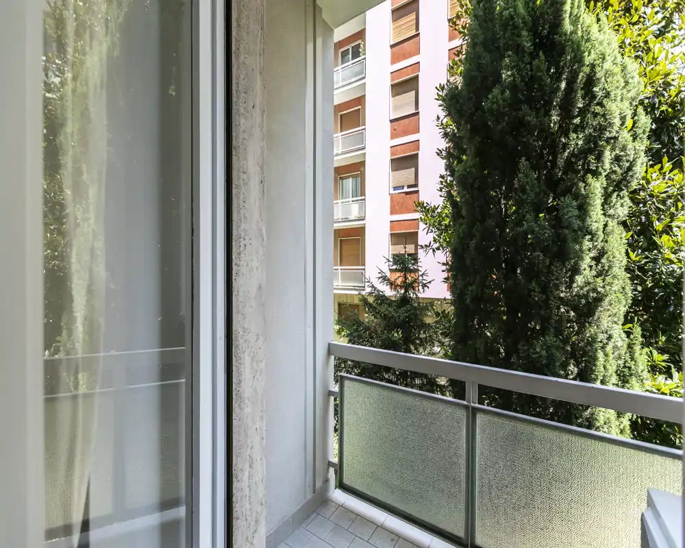 Balcón pequeño y soleado con barandilla de vidrio esmerilado, suelo de baldosas y vistas a árboles y fachadas de edificios cercanos — ideal para un café por la mañana.