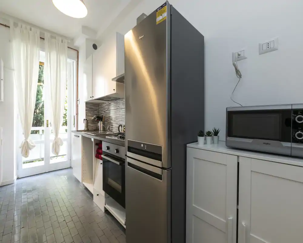 Cocina moderna tipo galería con frigorífico de acero inoxidable, horno y microondas; luz natural desde las puertas y acabado limpio y contemporáneo.