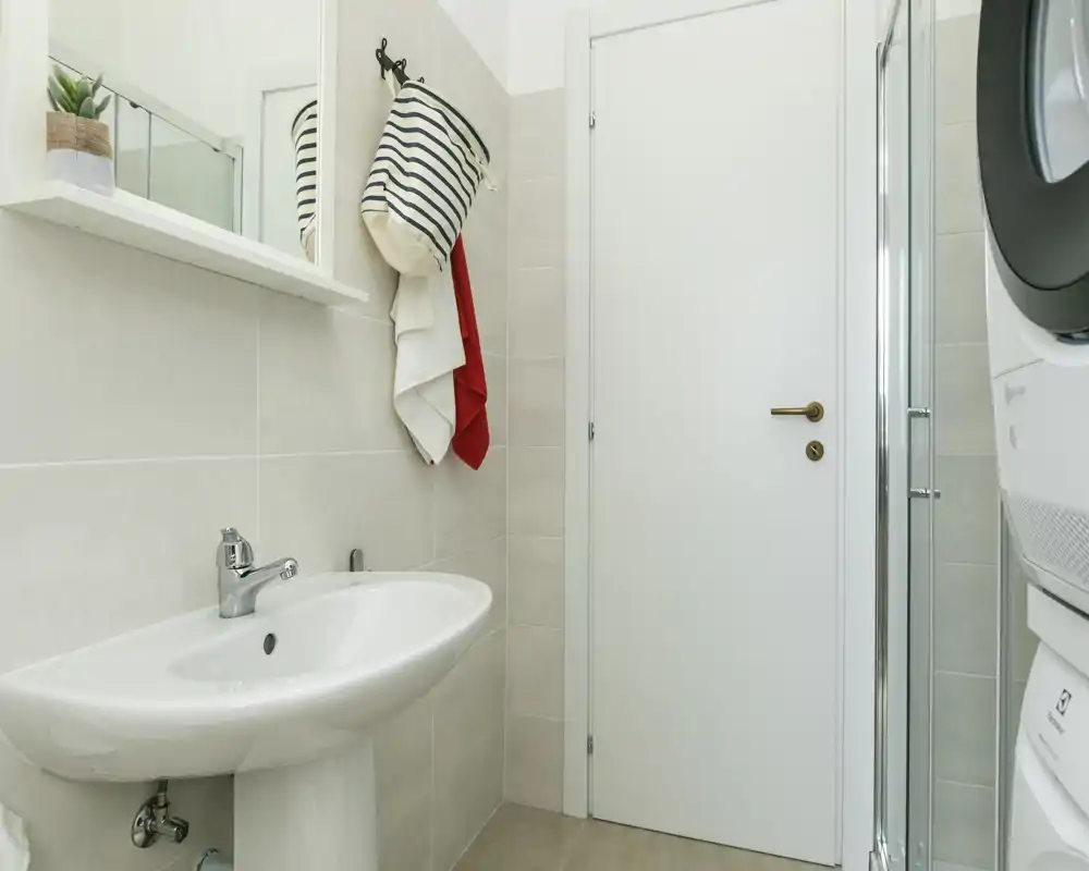 Baño moderno y limpio con lavabo de pedestal, espejo-armario, ganchos para toallas y ducha compacta; azulejos neutros y luminosos que aportan sensación de frescura.