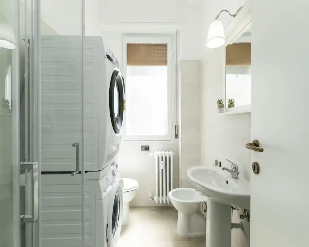 Baño luminoso y moderno con lavadora y secadora apiladas, lavabo de pie, bidé y ducha. Estilo limpio y aireado con luz natural que entra por la ventana.