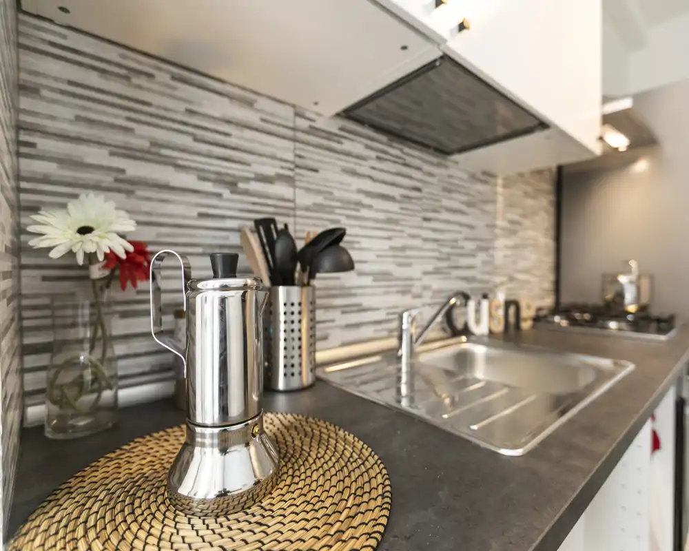 Rincón de cocina moderno y compacto con fregadero de acero inoxidable, cocina a gas y salpicadero de azulejos; detalles decorativos como cafetera, soporte de utensilios y un pequeño jarrón con flores.