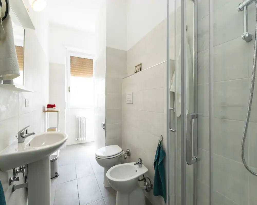 Baño moderno y luminoso con lavabo, inodoro, bidé y ducha con mampara de cristal; azulejos neutros y luz natural por la ventana que crean un ambiente limpio y fresco.