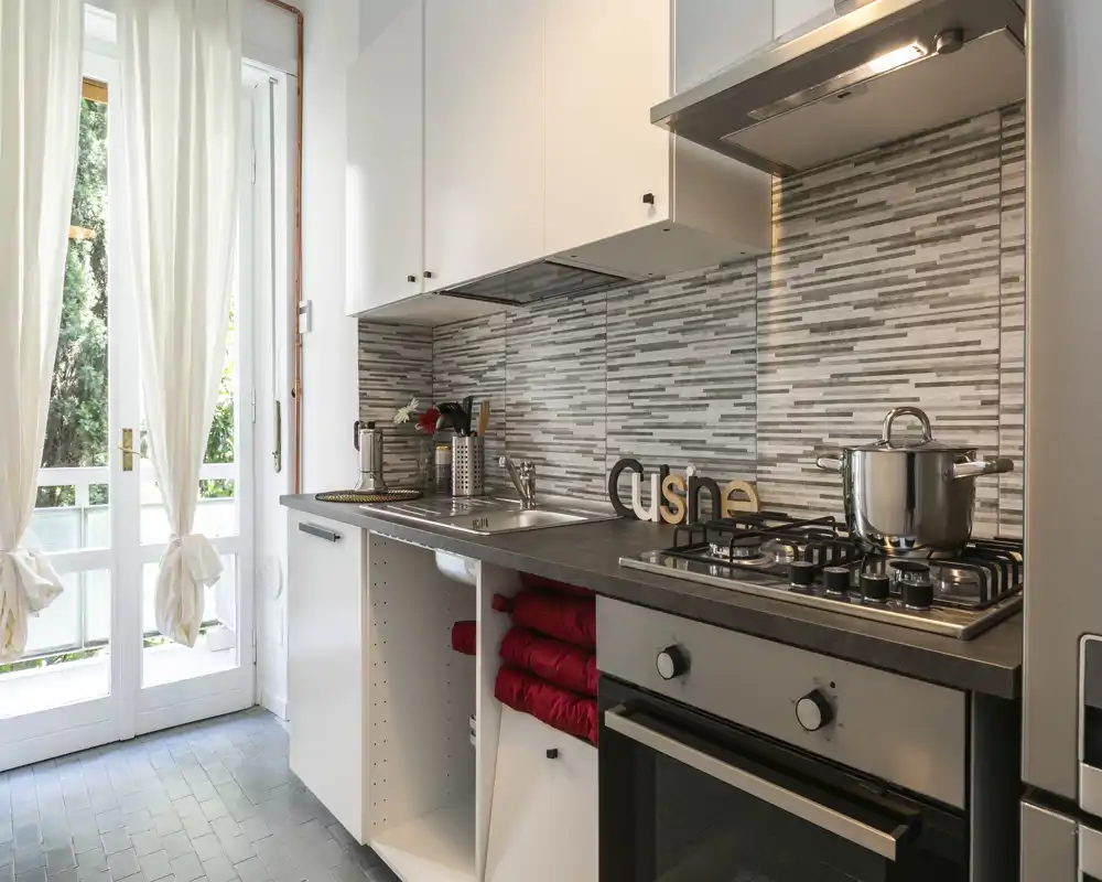 Cocina moderna y compacta con cocina de gas, horno y fregadero de acero inoxidable, luz natural desde puertas ventanas y un salpicadero de azulejos decorativo; toallas rojas dobladas y pequeños detalles crean un ambiente acogedor.