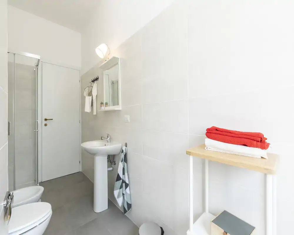 Baño moderno y luminoso con ducha, lavabo de pie, bidé y estantería con toallas plegadas. Baldosas neutras y decoración mínima que ofrecen una apariencia limpia y agradable.