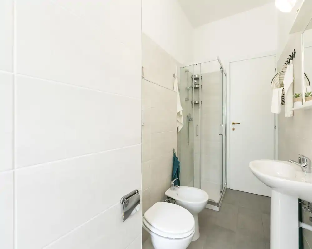 Baño moderno y luminoso con ducha, inodoro, bidé y lavabo de pedestal; azulejos neutros y líneas limpias para un aspecto fresco y minimalista.