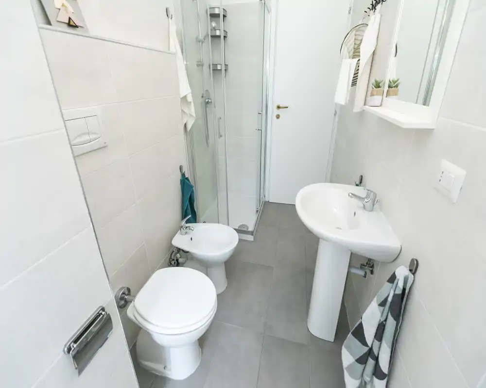 Baño pequeño y moderno con inodoro, bidé, lavabo con pedestal y ducha esquina de cristal. Azulejos neutros y accesorios sencillos que ofrecen un aspecto limpio y funcional.