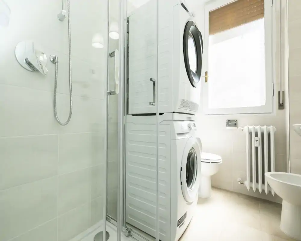 Baño moderno y compacto con ducha de cristal, lavadora y secadora apiladas, inodoro y bidé junto a una ventana luminosa.