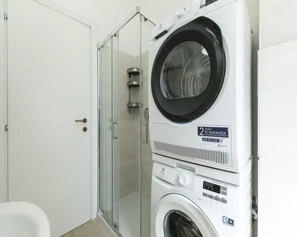 Baño compacto con lavadora y secadora apiladas junto a una ducha con mampara de cristal, azulejos neutros y luminosos y acabados modernos.