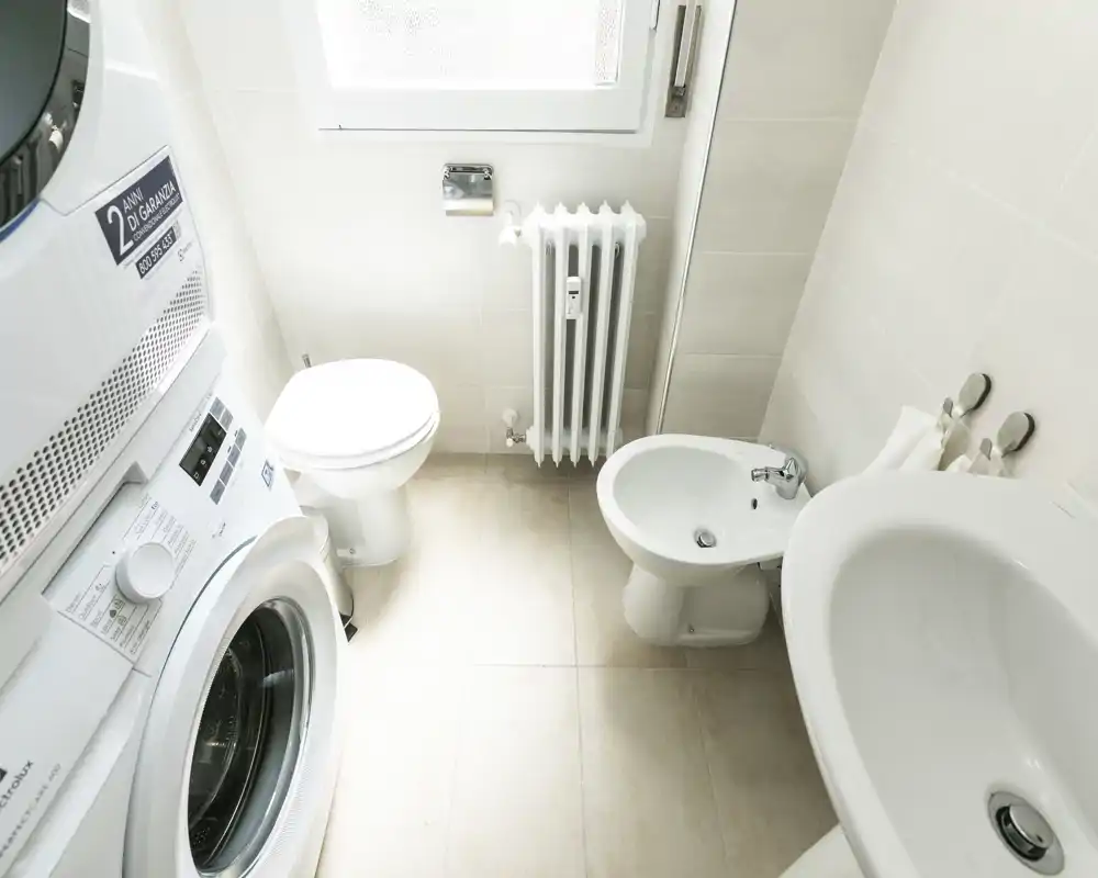 Baño compacto y luminoso con inodoro, bidé, lavabo y lavadora/secadora apiladas. Baldosas neutras y buena luz natural desde la ventana.