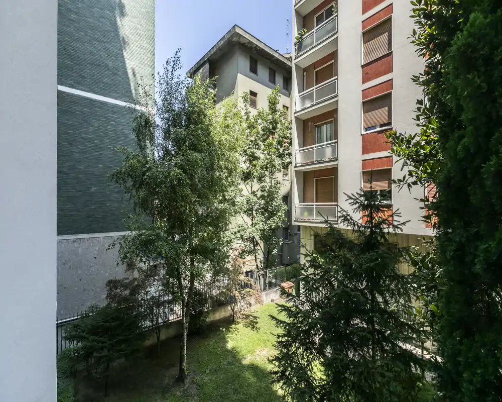 Pequeño jardín/patio privado entre edificios con árboles maduros, césped y balcones que dan al espacio verde.