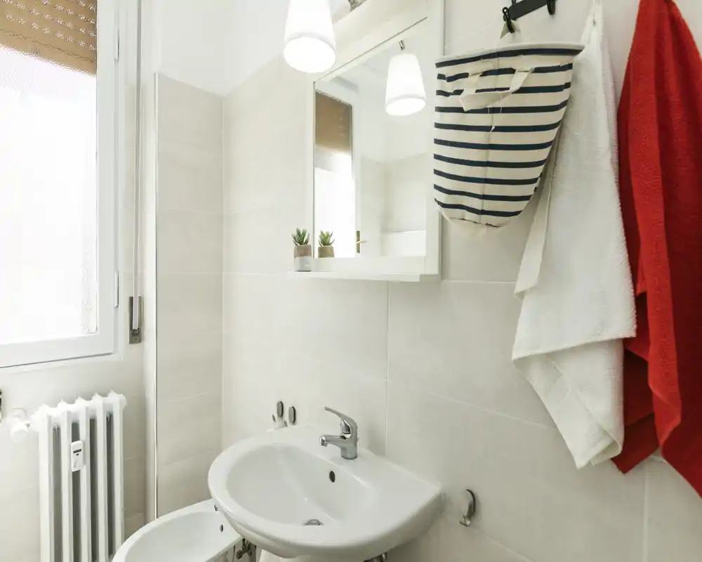 Baño luminoso y limpio con lavabo de pared, armario con espejo y luz natural procedente de una ventana. Toallas y un bolso colgante aportan funcionalidad.