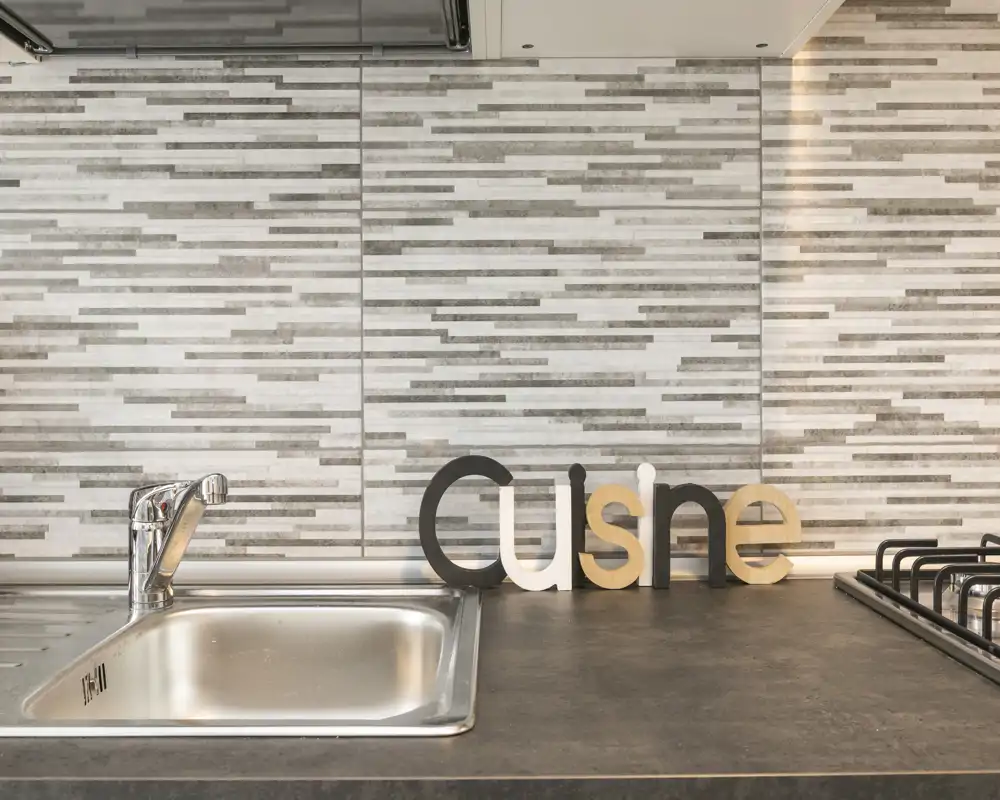 Encimera de cocina moderna con fregadero de acero inoxidable, placa de gas y letras decorativas de madera sobre un salpicadero en azulejos texturizados — luminosa, ordenada y preparada para la foto.