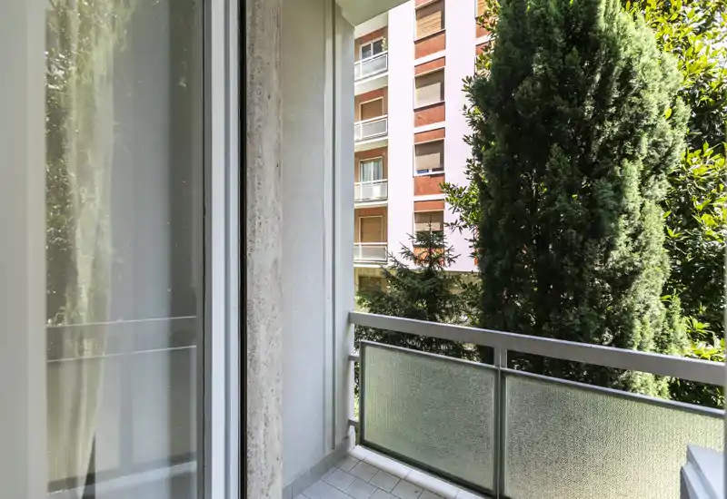 Sunny small balcony with frosted glass railing, tiled floor and views of neighboring trees and apartment façades — ideal for a morning coffee.